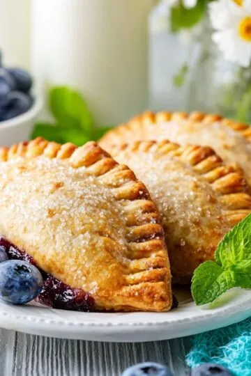 Three golden-brown, baked blueberry hand pies sprinkled with coarse sugar rest on a white plate garnished with fresh blueberries and mint.
