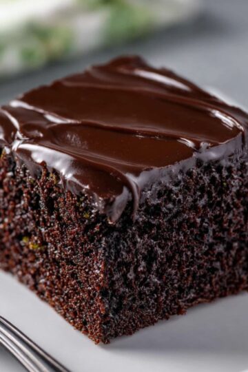 A thick slice of dark chocolate zucchini cake topped with rich chocolate frosting rests on a white dessert plate.