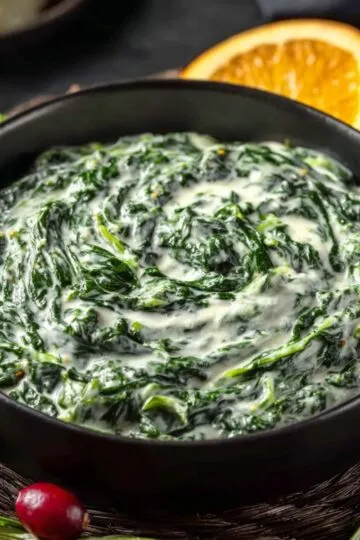 A dark serving bowl filled with rich, hot creamed spinach resting on a woven placemat next to fresh cranberries and an orange slice.