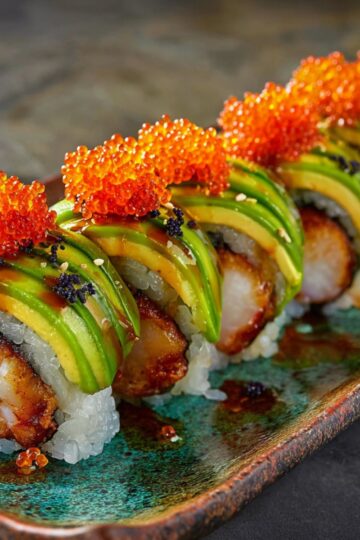 A plated Dragon Roll Sushi features a crispy shrimp center topped with overlapping avocado slices and sesame seeds.