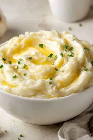 A large serving bowl filled with homemade mashed potatoes, garnished with a deep pool of melted butter and chopped green chives.