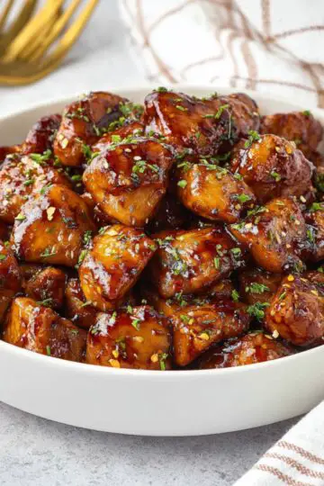 A bowl overflowing with glossy, caramelized honey garlic chicken bites garnished with fresh green parsley and red chili flakes.