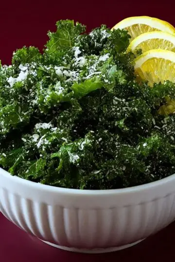 A white fluted bowl filled with bright green kale salad, heavily garnished with grated cheese and fresh lemon wedges against a dark background.