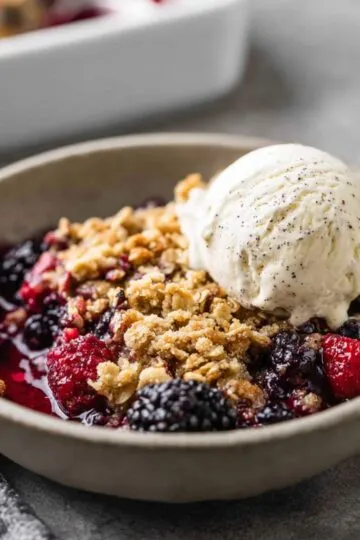 A bowl of warm mixed berry crisp with frozen berries features a golden oat crust and a melting scoop of vanilla ice cream.