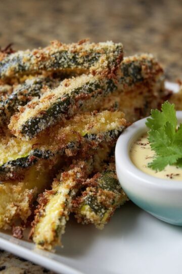 A white plate piled high with golden, crispy parmesan zucchini fries served next to a small bowl of creamy dipping sauce.