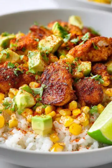 A hearty bowl filled with rice, seasoned chicken pieces, charred corn, crumbly cotija cheese, and a creamy dressing drizzle.